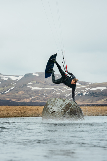 View of a kitesurfer performing a trick while touching a large rock in a river with snowy mountains in the background in Borgarbyggd Iceland. View of a kitesurfer performing a trick while touching a large rock in a river with snowy mountains in the background in Borgarbyggd Iceland.