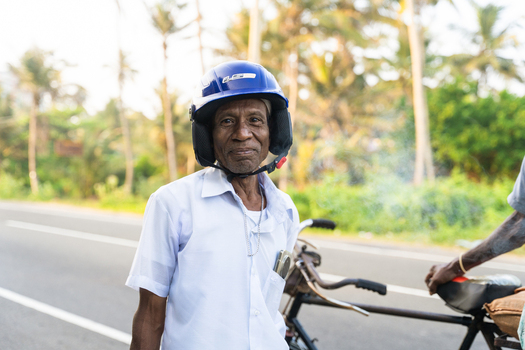 Sri Lanka 21 January 2026 View of an elderly man wearing a blue motorcycle helmet and white shirt standing by a bicycle on a road lined with palm trees.