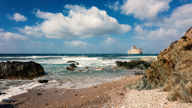 View of Scoglio Pan di zucchero sea stack from the rocky shore of Masua beach with crashing waves and white clouds in a blue sky in Masua Sardegna Italy. View of Scoglio Pan di zucchero sea stack from the rocky shore of Masua beach with crashing waves and white clouds in a blue sky in Masua Sardegna Italy.