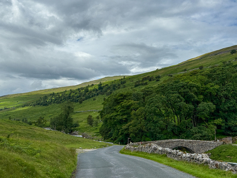 View of the Yorkshire countryside with a winding asphalt road leading past a historic stone bridge and lush green hills Hawes England United Kingdom. View of the Yorkshire countryside with a winding asphalt road leading past a historic stone bridge and lush green hills Hawes England United Kingdom.