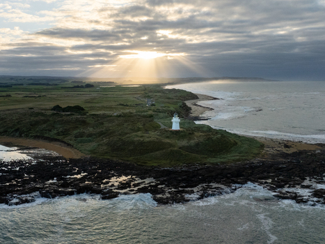 Aerial view of the lighthouse standing guard as the sun pierces through the clouds above the rugged coastline Otara Southland Region New Zealand.