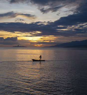 Aerial view of a lone fisherman in a small wooden boat on the calm sea during a vibrant sunset with distant mountains under a dramatic cloudy sky in Flores Indonesia.