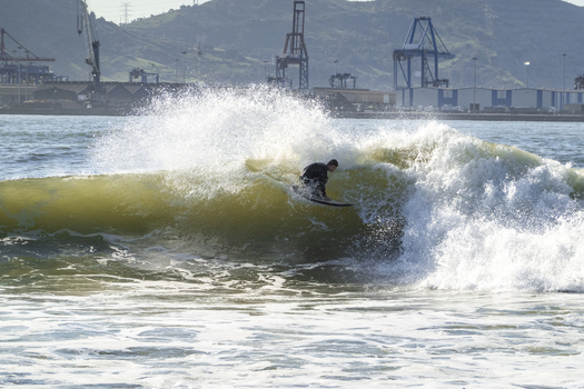 View of a surfer riding a breaking wave with white spray against an industrial port backdrop with cranes and hills at Ereaga Beach Algorta Euskadi Spain.