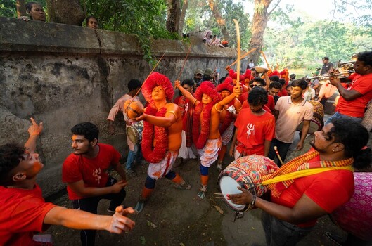 Munshiganj Bangladesh 13 April 2025 View of a procession red and orange figures celebrating with drums against a grey wall and green trees. Munshiganj Bangladesh 13 April 2025 View of a procession red and orange figures celebrating with drums against a grey wall and green trees.