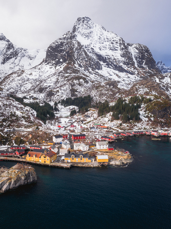 Aerial view of vibrant red and yellow buildings clinging to the rugged coastline beneath snow dusted mountains a striking contrast against the deep blue fjord Nusfjord Nordland Norway.