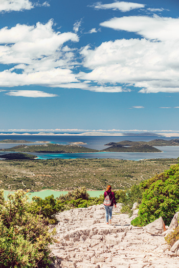 View of a woman walking on a rocky path towards Lake Vrana with islands and blue water under a cloudy sky in Vrana Zadar County Croatia. View of a woman walking on a rocky path towards Lake Vrana with islands and blue water under a cloudy sky in Vrana Zadar County Croatia.