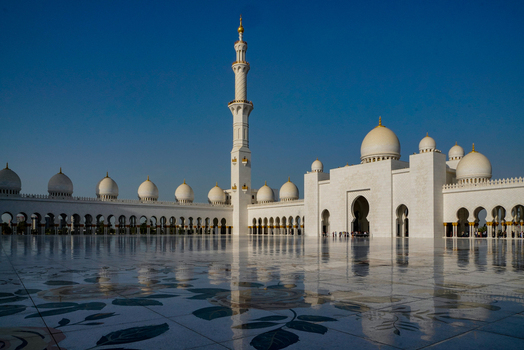 View of the Sheikh Zayed Grand Mosque featuring white marble domes a tall minaret and a reflective floral courtyard under a clear blue sky in Abu Dhabi United Arab Emirates. View of the Sheikh Zayed Grand Mosque featuring white marble domes a tall minaret and a reflective floral courtyard under a clear blue sky in Abu Dhabi United Arab Emirates.