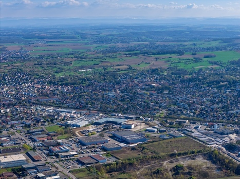 Aerial view of a sprawling cityscape where buildings and industrial structures meet verdant fields under a vast sky Sausheim Grand Est France.