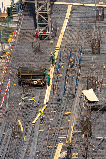 Aerial view of workers in safety gear walking on a steel rebar grid at a construction site with building materials and crane base in Dubai Dubai United Arab Emirates. Aerial view of workers in safety gear walking on a steel rebar grid at a construction site with building materials and crane base in Dubai Dubai United Arab Emirates.