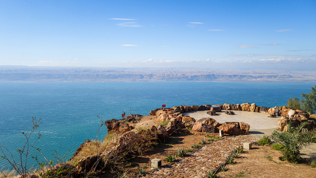 View of the Dead Sea from a rocky cliffside overlook with a stone wall and paved terrace under a clear blue sky in Jordan. View of the Dead Sea from a rocky cliffside overlook with a stone wall and paved terrace under a clear blue sky in Jordan.