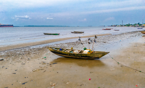 Aerial view of a wooden fishing boat resting on the sandy shore of Bonny Island with calm coastal waters and distant vessels in Bonny Town Rivers Nigeria. Aerial view of a wooden fishing boat resting on the sandy shore of Bonny Island with calm coastal waters and distant vessels in Bonny Town Rivers Nigeria.