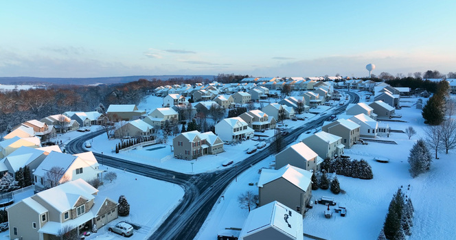 Aerial view of a suburban neighborhood blanketed in fresh snow with houses winding roads and a distant water tower under a clear winter sky in Manheim Pennsylvania United States.