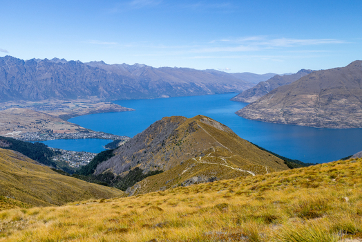 View of a stunning panorama featuring golden tussock grass a winding trail and the expansive blue lake against a backdrop of majestic mountains Ben Lomond Queenstown New Zealand. View of a stunning panorama featuring golden tussock grass a winding trail and the expansive blue lake against a backdrop of majestic mountains Ben Lomond Queenstown New Zealand.