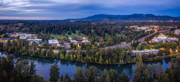 Aerial view of the Willamette River and downtown Eugene at twilight with riverfront development green spaces streets and illuminated buildings. Aerial view of the Willamette River and downtown Eugene at twilight with riverfront development green spaces streets and illuminated buildings.