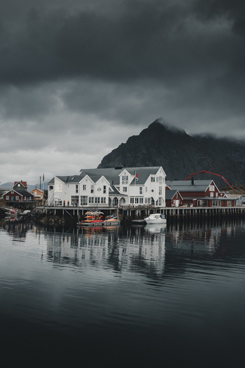 View of Henningsvaer fishing village with white and red wooden buildings on stilts calm water reflections and a dramatic mountain backdrop under dark clouds in Henningsvaer Nordland Norway. View of Henningsvaer fishing village with white and red wooden buildings on stilts calm water reflections and a dramatic mountain backdrop under dark clouds in Henningsvaer Nordland Norway.