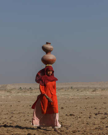 Yazman Punjab Pakistan 19 March 2010 View of a woman in traditional red clothing carrying stacked clay pots on her head while walking through the arid desert landscape near Chanan Pir.