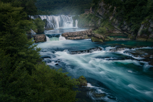 View of the Strbacki buk waterfall cascading over rocky tiers on the Una River with lush green forest.