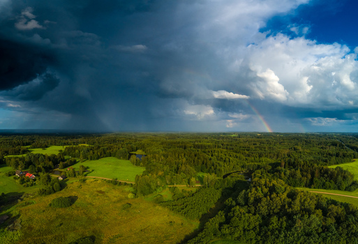 Aerial view of a lush green landscape with dense forests and fields under dramatic dark storm clouds and a vibrant rainbow in Latvia. Aerial view of a lush green landscape with dense forests and fields under dramatic dark storm clouds and a vibrant rainbow in Latvia.