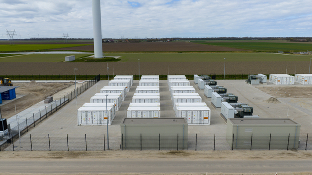 Aerial view of a battery energy storage system with rows of white containers and electrical equipment in a fenced facility near a wind turbine in Dronten Flevoland Netherlands.