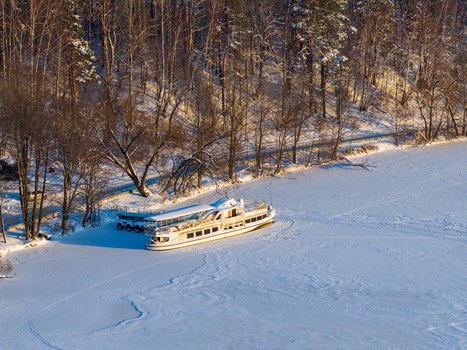 Aerial view of a white passenger boat docked on a frozen lake surrounded by snow covered ground and a dense forest in Trakai Vilnius County Lithuania. Aerial view of a white passenger boat docked on a frozen lake surrounded by snow covered ground and a dense forest in Trakai Vilnius County Lithuania.