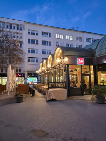 Hanover Germany 30 March 2026 View of an outdoor cafe area with illuminated arched glass structures and modern urban architecture in the background during twilight.