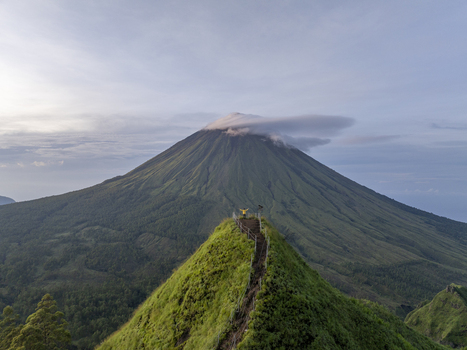 Aerial view of Mount Inerie with a person standing on a grassy ridge lush volcanic slopes and a cloud cap at the summit in Inerie Flores Indonesia. Aerial view of Mount Inerie with a person standing on a grassy ridge lush volcanic slopes and a cloud cap at the summit in Inerie Flores Indonesia.
