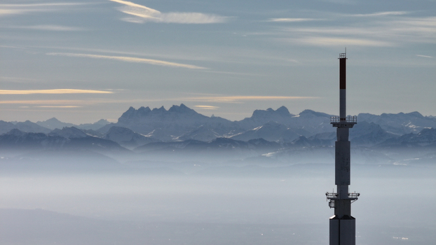 Aerial view of a soaring radio tower piercing through a sea of mist with snow capped mountains fading into the horizon Gex Auvergne Rhône Alpes France. Aerial view of a soaring radio tower piercing through a sea of mist with snow capped mountains fading into the horizon Gex Auvergne Rhône Alpes France.