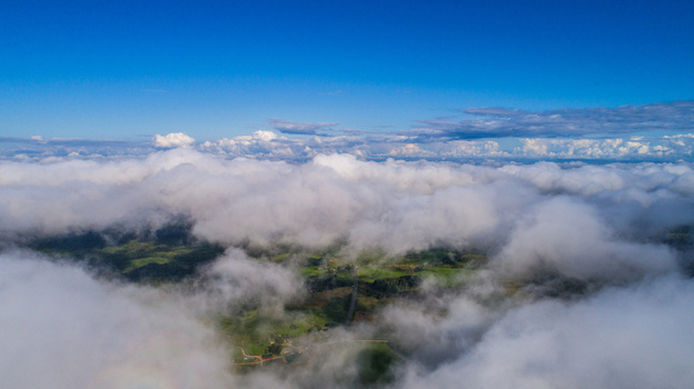 Aerial view of a landscape partially obscured by a thick layer of white clouds with green fields and a road visible below under a bright blue sky in Latvia. Aerial view of a landscape partially obscured by a thick layer of white clouds with green fields and a road visible below under a bright blue sky in Latvia.
