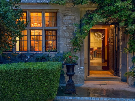 View of a historic stone manor house entrance with warm interior lighting through leaded windows an open doorway and lush garden greenery Hawes England United Kingdom. View of a historic stone manor house entrance with warm interior lighting through leaded windows an open doorway and lush garden greenery Hawes England United Kingdom.