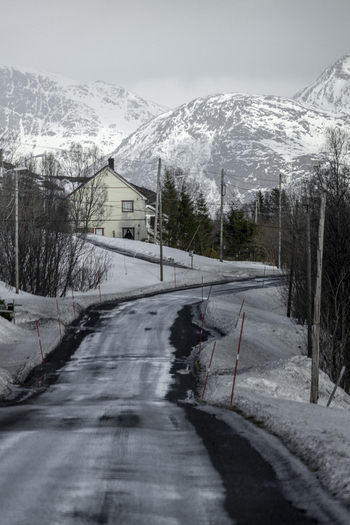 View of a winding icy road leading towards a white house and snow covered mountains under an overcast sky in Kvaloya Troms Norway. View of a winding icy road leading towards a white house and snow covered mountains under an overcast sky in Kvaloya Troms Norway.
