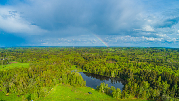 Aerial view of a serene lake surrounded by lush green forests and meadows under a dramatic sky with a faint rainbow in Latvia. Aerial view of a serene lake surrounded by lush green forests and meadows under a dramatic sky with a faint rainbow in Latvia.