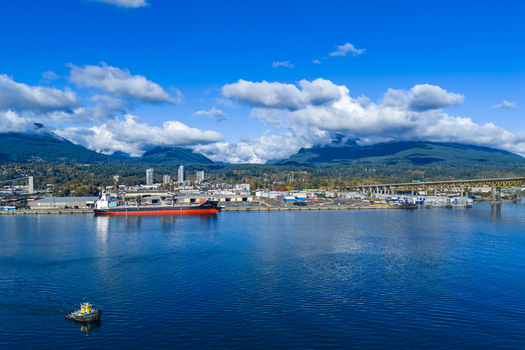 Aerial view of Vancouver Harbour featuring a large cargo ship at the industrial port a small tugboat in the water and mountains under a bright sky in Vancouver British Columbia Canada. Aerial view of Vancouver Harbour featuring a large cargo ship at the industrial port a small tugboat in the water and mountains under a bright sky in Vancouver British Columbia Canada.