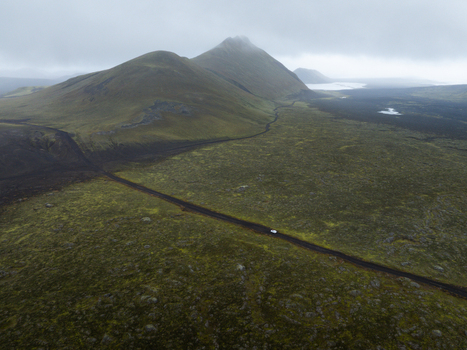 Aerial view of a lone car traverses a stark winding road through mossy highlands juxtaposed against dark volcanic earth under a misty sky Highlands Iceland.