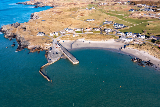 Aerial view of the harbour and sandy beach at Portnablagh with a stone pier extending into clear blue waters surrounded by coastal hills and houses in Portnablagh County Donegal Ireland. Aerial view of the harbour and sandy beach at Portnablagh with a stone pier extending into clear blue waters surrounded by coastal hills and houses in Portnablagh County Donegal Ireland.