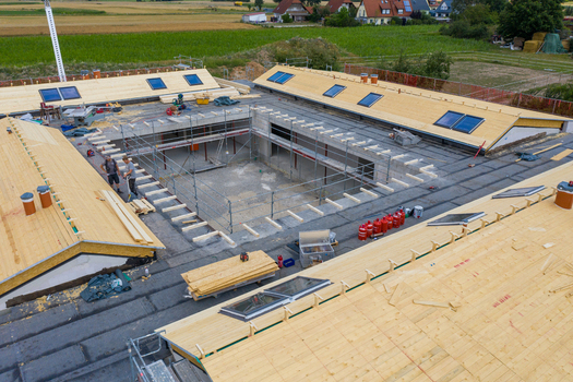 Igensdorf Germany 11 May 2024 Aerial view of a building under construction with wooden roof panels scaffolding and workers on site surrounded by green fields. Igensdorf Germany 11 May 2024 Aerial view of a building under construction with wooden roof panels scaffolding and workers on site surrounded by green fields.