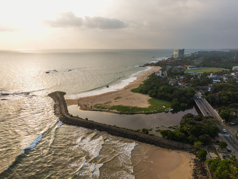 Aerial view of the coastline featuring a stone breakwater sandy beach and a river mouth near a bridge and lush tropical trees in Galle Southern Province Sri Lanka.