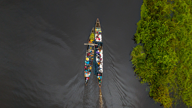 Aerial view of two wooden canoes filled with people and goods navigating the dark waters of a river beside lush green vegetation in Itu Akwa Ibom Nigeria. Aerial view of two wooden canoes filled with people and goods navigating the dark waters of a river beside lush green vegetation in Itu Akwa Ibom Nigeria.