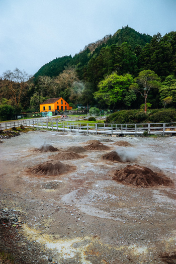 View of steaming fumaroles dotting the volcanic landscape contrasting with the vibrant yellow house nestled against the lush green hillside Furnas Azores Portugal.