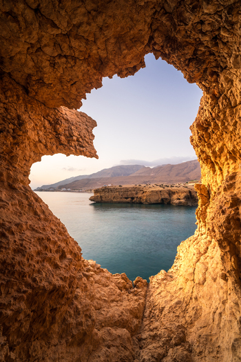 View of the turquoise sea and rugged mountains framed by a natural rocky cave opening in Oman. View of the turquoise sea and rugged mountains framed by a natural rocky cave opening in Oman.