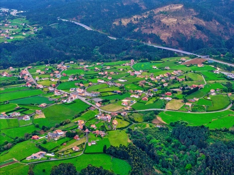 Aerial view of a rural village with houses scattered across vibrant green fields and forested hills near a highway viaduct in Cudillero Asturias Spain.