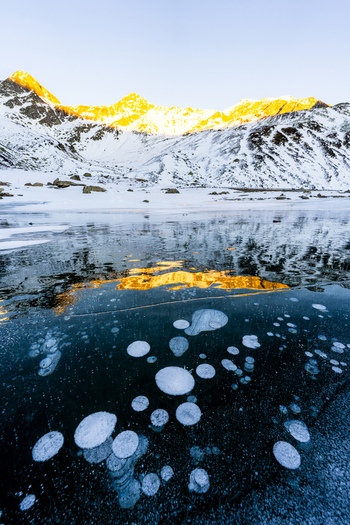 View of methane bubbles trapped in a frozen lake with snow covered mountains at sunset in Madesimo Lombardy Italy. View of methane bubbles trapped in a frozen lake with snow covered mountains at sunset in Madesimo Lombardy Italy.