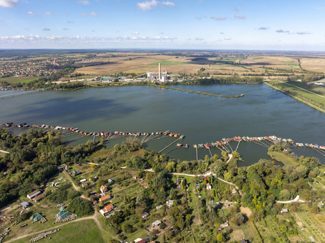 Aerial view of Bokodi Floating Village featuring colorful wooden houses on stilts over the lake lush greenery and an industrial plant in the distance in Oroszlany Hungary. Aerial view of Bokodi Floating Village featuring colorful wooden houses on stilts over the lake lush greenery and an industrial plant in the distance in Oroszlany Hungary.