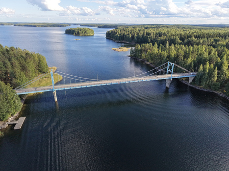 Aerial view of Potkusalmen silta Bridge spanning the calm waters of a lake surrounded by dense pine forests under a clear blue sky in Savonlinna Etela Savo Finland.