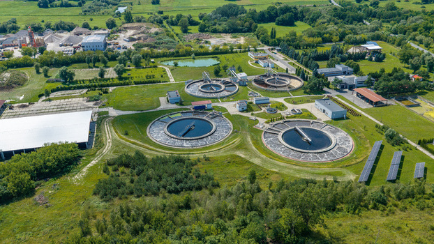 Aerial view of a wastewater treatment plant with circular sedimentation tanks industrial buildings and solar panels surrounded by green fields and trees in Poland.