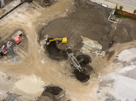 Aerial view of a yellow excavator digging into a large pile of dark earth next to industrial machinery and gravel mounds in San Giovanni Valdarno Tuscany Italy.