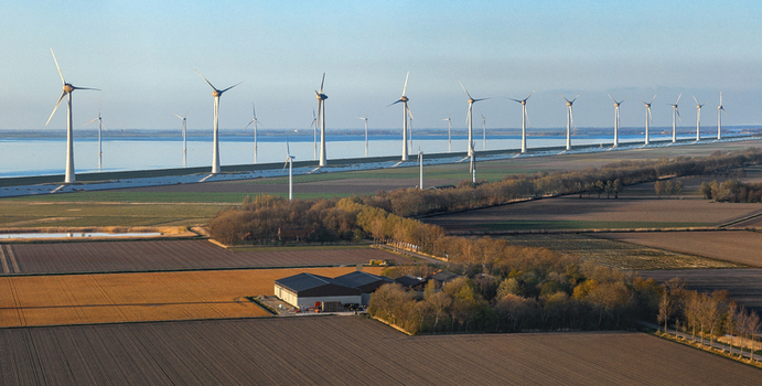 Aerial view of a row of wind turbines along a dike bordering agricultural fields and water under a clear sky Netherlands.
