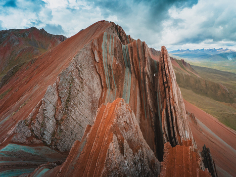 Aerial view of the striking jagged peaks of Pallay Punchu Rainbow Mountain with its distinct red and turquoise mineral layers under a dramatic cloudy sky in Layo Cuzco Peru.