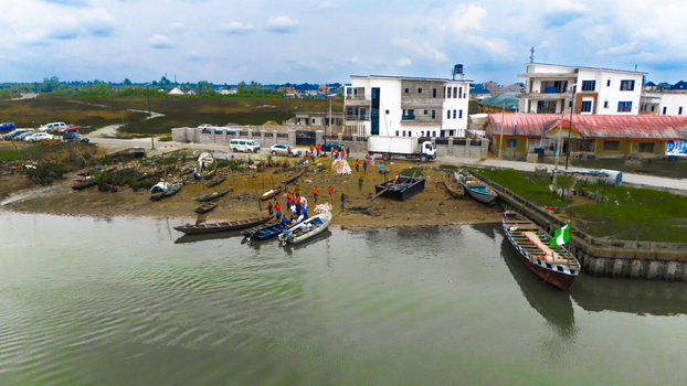 Aerial view of a riverbank in Rivers Nigeria featuring wooden boats docked on the muddy shore people gathering near the water and buildings in the background.