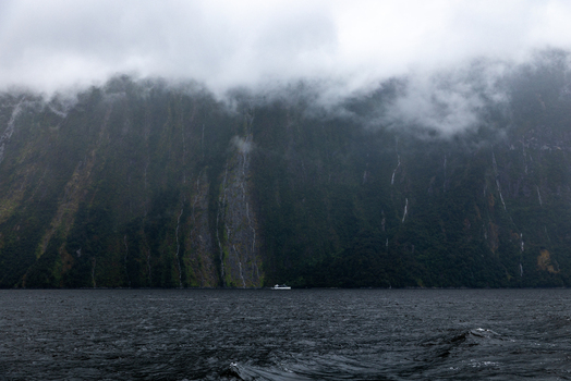 View of mist shrouded cliffs cascading into the dark waters where a small boat navigates the fjord Milford Sound Southland Region New Zealand.