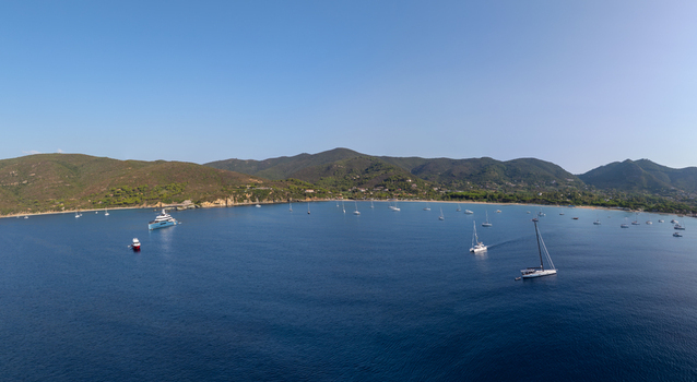 Aerial view of Lacona Beach with yachts and sailboats anchored in the clear blue bay surrounded by green hills under a clear sky in Lacona Tuscany Italy.